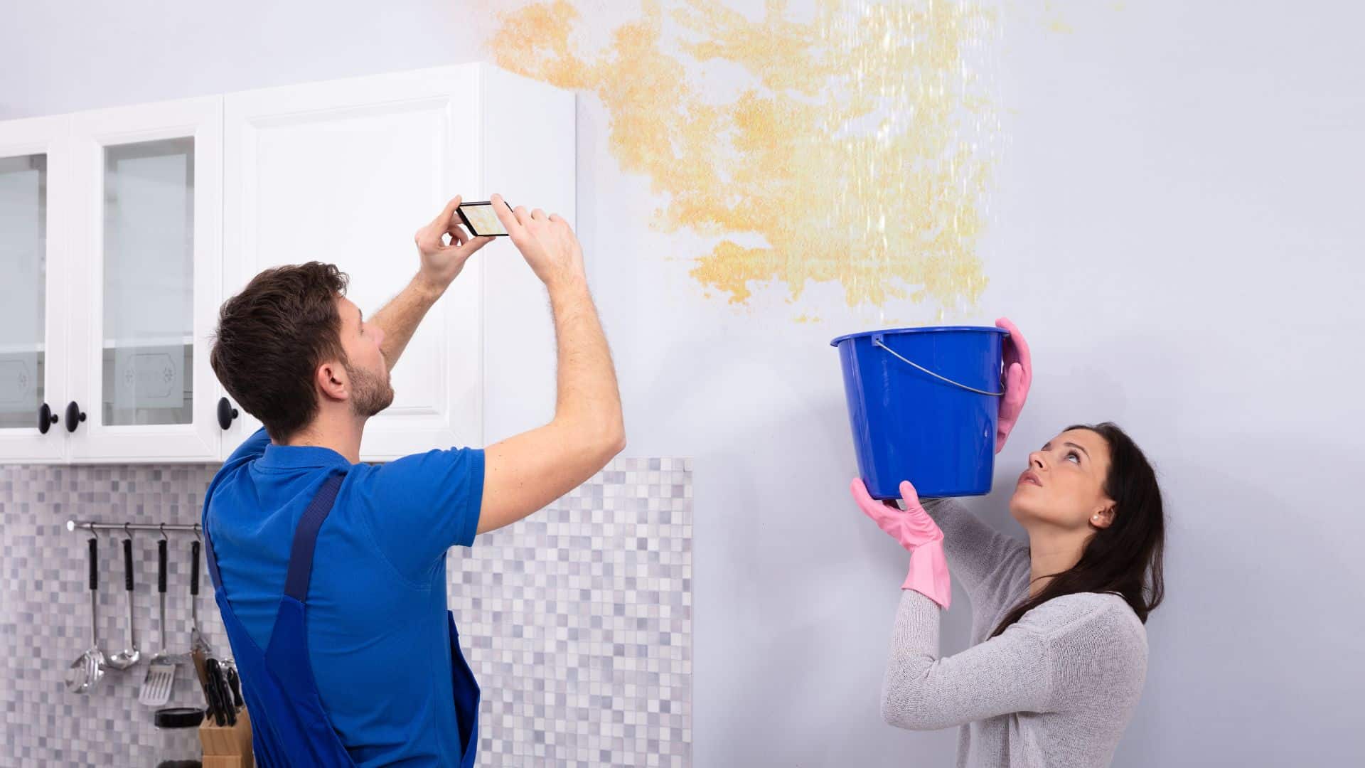 Man taking photos of a water-damaged kitchen ceiling while a woman holds a bucket under the leak.